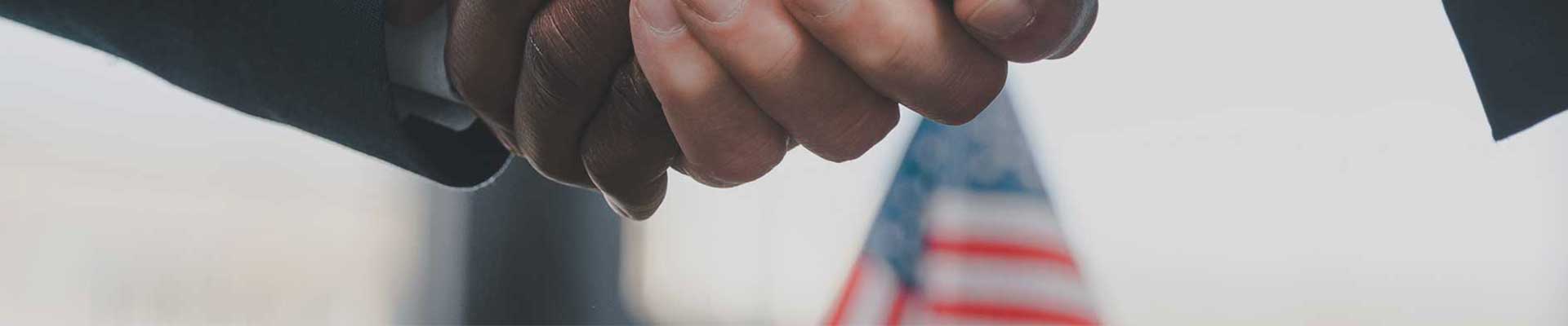 Two individuals clasping hands with an out-of-focus american flag in the background.